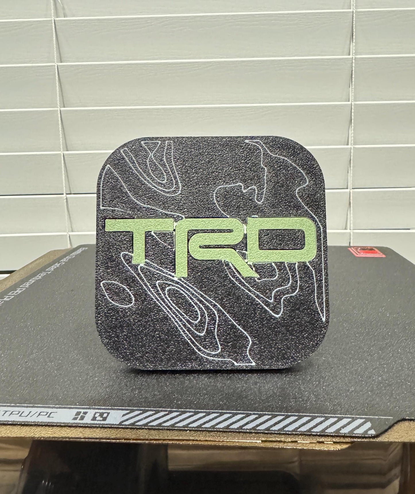 May include: Black square-shaped emblem with a white topographic pattern and a green "TRD" logo.
